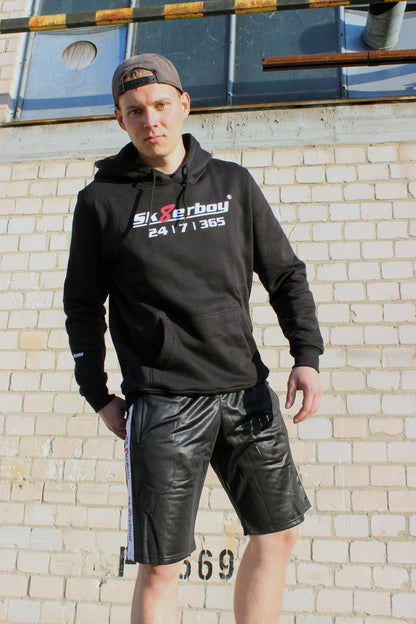 boy traegt einen sk8erboy kapuzen pullover mit einem aufdruck sk8erboy 365 7 24 mit einem eindeutigen slogan ebenso sk8erboy acht 8 in rot mit weissem schriftzug auf der brust und dem aermel der boy traegt eine cap im hintergrund ein lost place