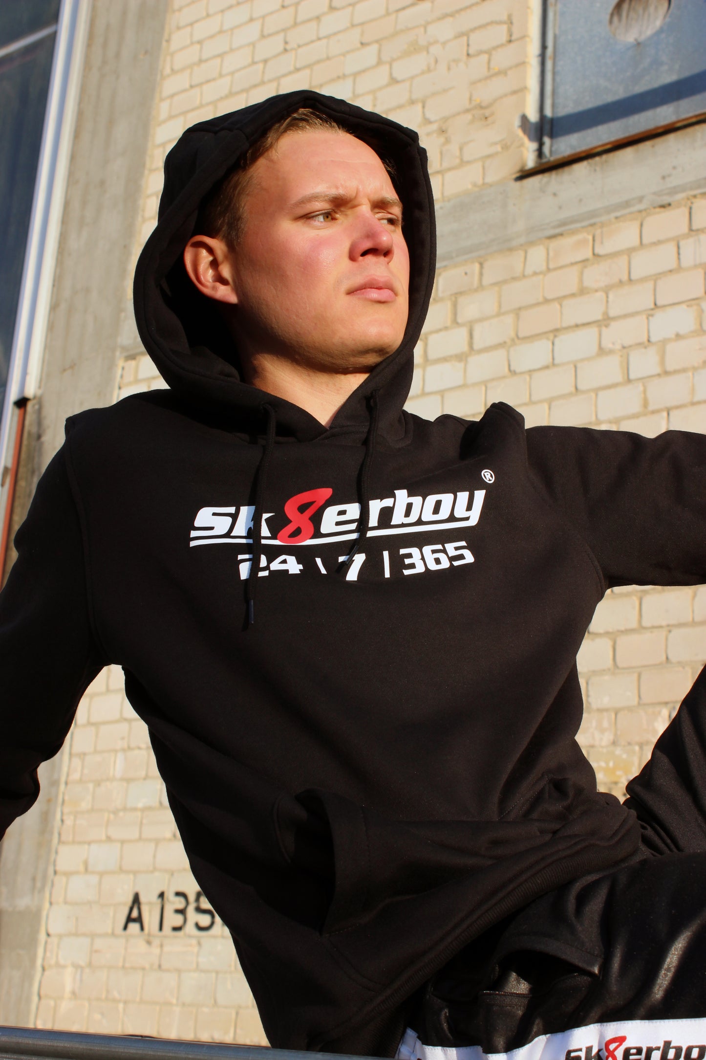 boy traegt einen sk8erboy kapuzen pullover mit einem aufdruck sk8erboy 365 7 24 mit einem eindeutigen slogan ebenso sk8erboy acht 8 in rot mit weissem schriftzug auf der brust und dem aermel der boy traegt eine cap im hintergrund ein lost place er geniesst die sonne