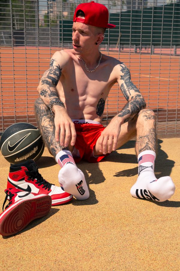 junger sportlicher gay mit trainiertem oberkoerper in nahaufnahme mit tattoos an beinen und armen zieht sich die weissen socken mit usa flagge neben seinen roten nike turnschuhen auf einem basketball feld traegt eine glanz boxer shorts unterhose in rot und basecap