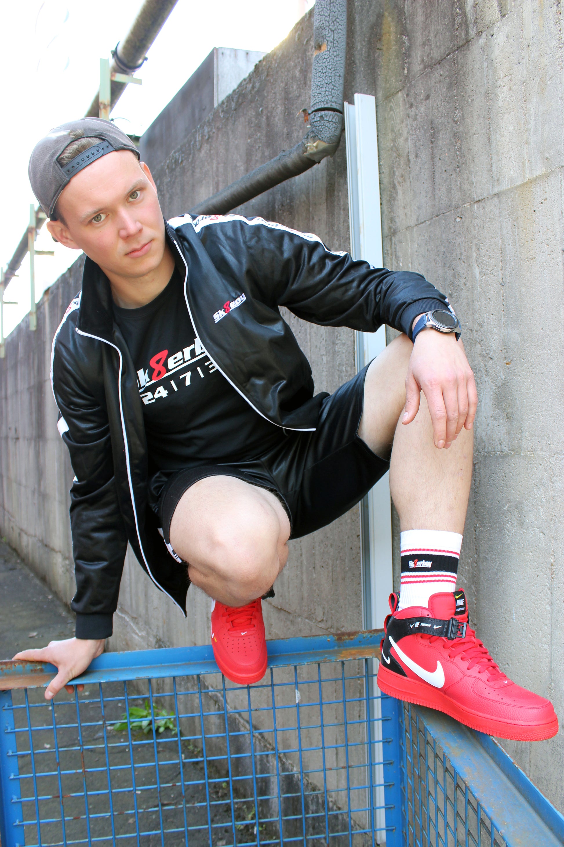 ein boy kerl bengel traegt ein sk8erboy t-shirt shirt in schwarz baumwolle cotton mit einem sk8erboy aufdruck mit weisser schrift und 8 acht in rot mit 24 7 365 fetter aufdruck weiter traegt er eine mund nasen bedenkung maske sk8erboy mit aufdruck dann hat er auf dem kopf eine basecap baseball seine graue jogger spant in der hose von nike air max und im hintergrund ein lost place alte fabrik