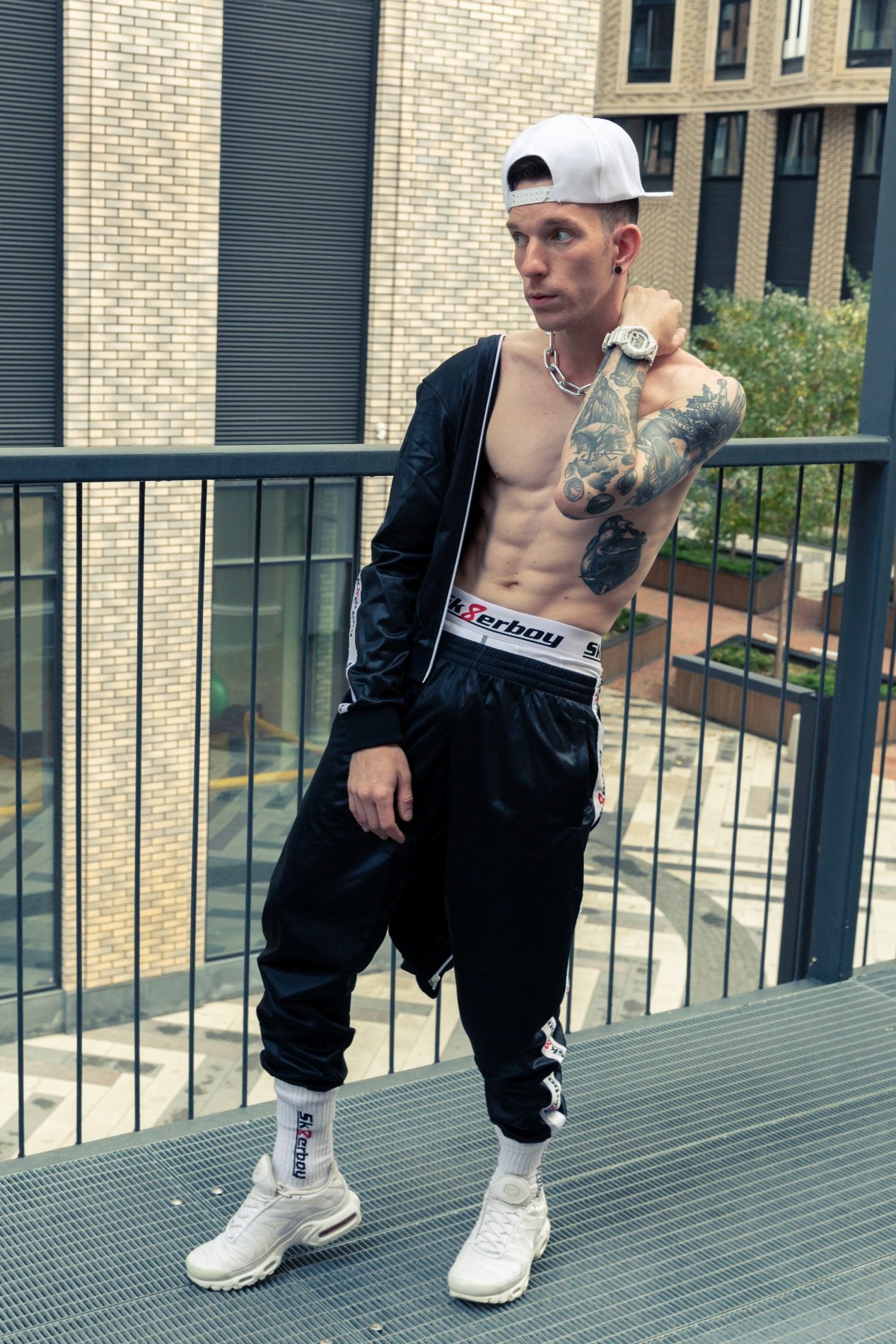 sportlicher junge trägt einen glanz trainingsanzug in schwarz von und passende crew socken mit weisser boxershort von sk8erboy in einer stadt auf der terrasse mit weissen nike tn sneakern und kappe