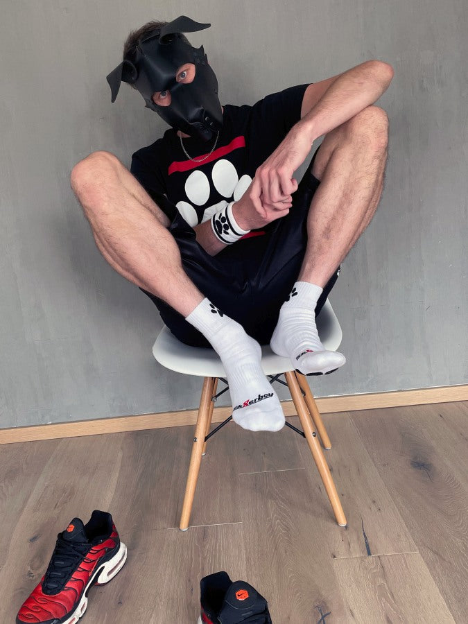 junger gay doggy traegt weisse sk8erboy puppy short crew socks mit hunde pfoten in schwarz und dog paw t-shirt mit hundemaske zu seinen roten nike tn auf einem stuhl sitzend vor einer wand
